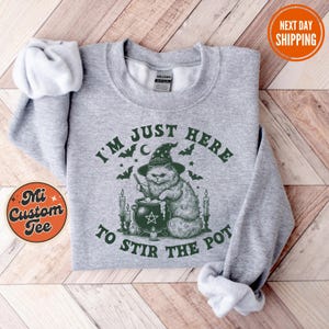 Puede incluir: Sudadera gris claro con el texto verde "I'M JUST HERE TO STIR THE POT". El diseño presenta un gato con sombrero de bruja revolviendo un caldero. Murciélagos y velas rodean al gato. La sudadera tiene mangas largas.