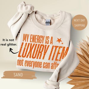 Puede incluir: Una sudadera de color arena con el texto "MY ENERGY IS A LUXURY ITEM not everyone can afford" en naranja. La sudadera tiene cuello redondo y mangas largas. Las palabras "It is not real glitter" también están en la imagen.