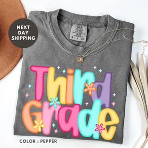 a gray t-shirt with a colorful design that says "Third Grade" and includes flowers. The t-shirt is placed on a white surface, and there is a brown hat and some leaves in the background.