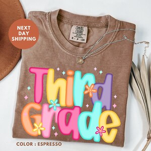 a brown t-shirt with a colorful design that says "Third Grade" and includes flowers. The t-shirt is placed on a white surface, and there is a brown hat and some leaves nearby.
