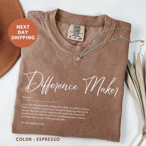 May include: A brown t-shirt with the text "Difference Maker" in white. The text is defined as a noun and describes a person who inspires others to be their best. The shirt is available in the color espresso.