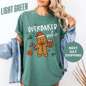 May include: Light green t-shirt with the text "OVERBAKED & OVER IT!" and a gingerbread man graphic wearing a Santa hat. The graphic includes a candy cane, gift, and a mug of marshmallows. The shirt is a relaxed fit.