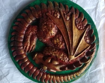 Puede incluir: Una placa de pared redonda de cerámica verde y dorada con un dragón esculpido detallado con escamas rojas y doradas. El dragón está enroscado con las alas extendidas.