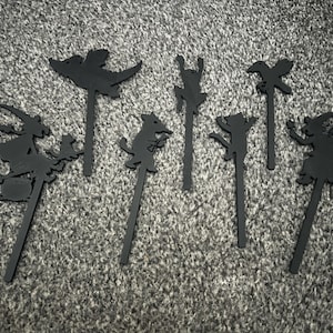Könnte beinhalten: Ein Set aus sechs flachen, schwarzen Silhouetten-Kuchentoppern mit Halloween-Motiven, darunter eine Hexe auf einem Besen, ein fliegender Drache, eine Katze, eine Spinne, eine Krähe und eine zweite Hexenfigur, jeweils an einem dünnen Stab.