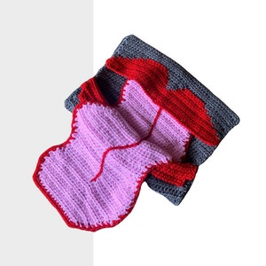 Puede incluir: Una manta de ganchillo en forma de corazón en rosa, rojo y gris. La manta está hecha de hilo suave y tiene un diseño único.