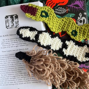 May include: Crocheted snake bookmarks in green, black, and white, with a tan tassel. The bookmarks are placed on an open book with text and a decorative cover. The snake bookmarks are handmade and add a unique touch to reading.