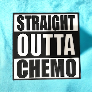May include: A black and white sign with the words "STRAIGHT OUTTA CHEMO" in bold, capital letters. The text is arranged in three lines, with "OUTTA" in the center. The sign is rectangular and sits on a light blue surface.