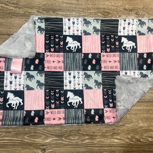 May include: A pink and black patchwork baby blanket with a gray fleece backing. The blanket features a repeating pattern of horses, arrows, and the text "Wild and Free".