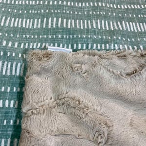 May include: A close-up of a soft, plush, taupe-colored blanket. The blanket has a textured, faux fur appearance. The background is a green and white patterned fabric. The blanket is folded over the patterned fabric.