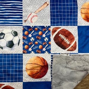 May include: A patchwork sports-themed blanket featuring squares with a baseball bat and ball, a soccer ball, a football, and a basketball. The blanket has blue and gray squares with a soft, gray, plush backing.