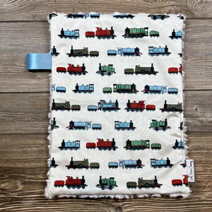 May include: A white baby blanket with a repeating pattern of colorful toy trains. The trains are in various colors, including red, green, blue, and brown. The blanket has a soft, fluffy border and a light blue ribbon attached to the side.