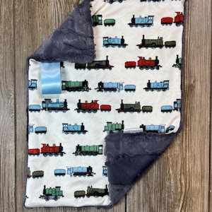 May include: A white baby blanket with a gray minky backing. The blanket features a repeating pattern of colorful toy trains on a white background.