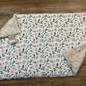 May include: A soft, white and beige baby blanket with a floral pattern featuring peach-colored roses and green leaves. The blanket is made of two layers, one side is a soft, plush fabric and the other side is a cotton fabric with a floral print.