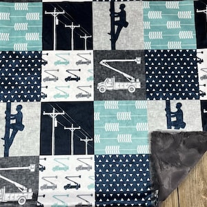 May include: A blue and white baby blanket with a patchwork design featuring images of utility trucks, power lines, and a lineman climbing a pole. The blanket has a soft, gray fleece backing.