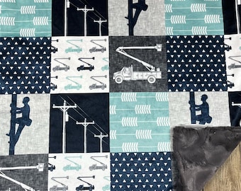May include: A blue and white baby blanket with a patchwork design featuring images of utility trucks, power lines, and a lineman climbing a pole. The blanket has a soft, gray fleece backing.
