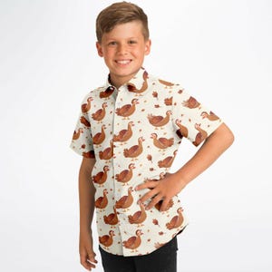 Puede incluir: Una camisa blanca de manga corta con botones y un estampado de pavos marrones y hojas naranjas.