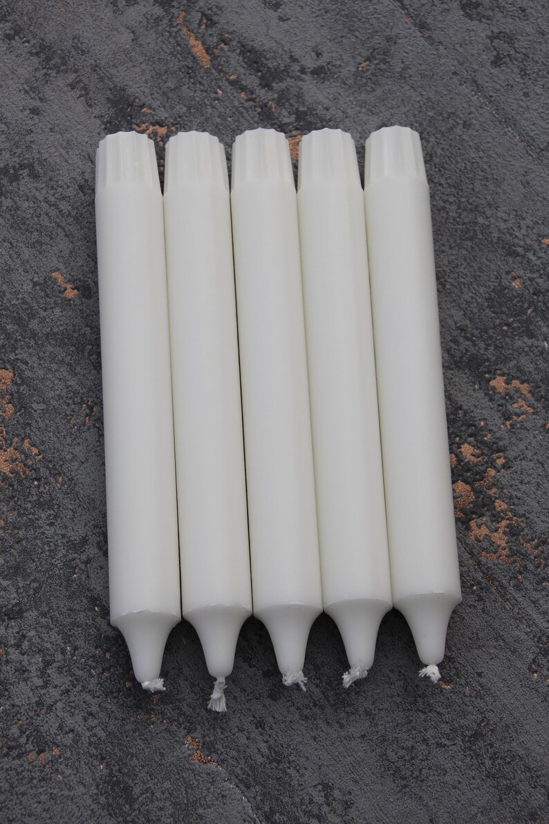 May include: Five white taper candles with a textured top and a pointed bottom. The candles are arranged in a row on a gray textured surface.