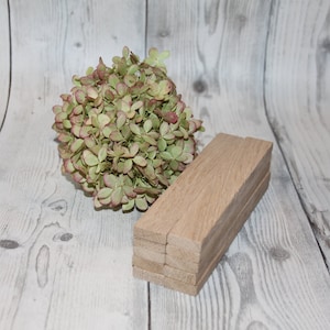 May include: A stack of six wooden rectangular blocks, each with a natural wood finish. The blocks are arranged in a staggered pattern, with the top block slightly offset from the bottom block. The blocks are sitting on a white wooden surface. A bouquet of dried hydrangeas with green leaves and pink accents is in the background.