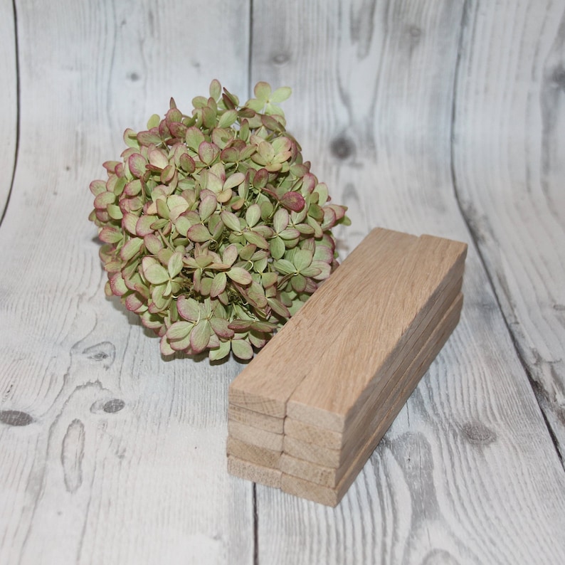 May include: A stack of six wooden rectangular blocks, each with a natural wood finish. The blocks are arranged in a staggered pattern, with the top block slightly offset from the bottom block. The blocks are sitting on a white wooden surface. A bouquet of dried hydrangeas with green leaves and pink accents is in the background.