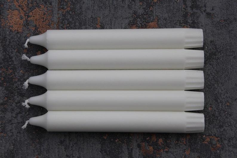 May include: Five white taper candles with a fluted design at the top. The candles are arranged in a stack.