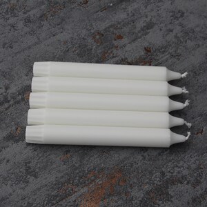 May include: A stack of six white taper candles with a pointed tip. The candles are arranged in a row on a gray textured surface.