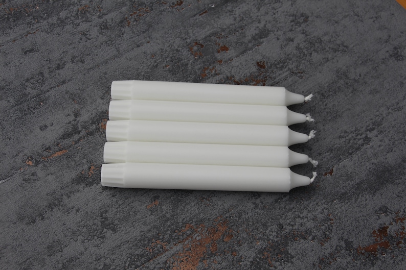 May include: A stack of six white taper candles with a pointed tip. The candles are arranged in a row on a gray textured surface.