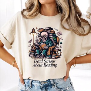 Könnte beinhalten: Ein cremefarbenes T-Shirt mit einer Grafik eines Skeletts mit Brille, das in einem Sessel sitzt und ein Buch liest, umgeben von Bücherstapeln, einer Lampe und einer Kaffeetasse, mit dem darunter gedruckten Text "Dead Serious About Reading".