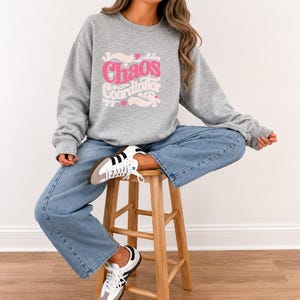 May include: A heather grey long-sleeve crewneck sweatshirt featuring the text "Chaos Coordinator" in a pink and white retro-style font, accented with small pink hearts and decorative swirls, paired with light blue denim jeans and white trainers.