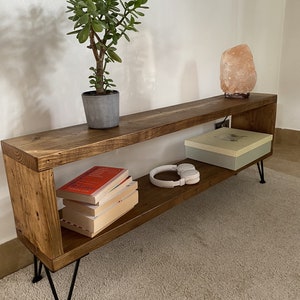 May include: A rustic wooden shelf with two shelves and black hairpin legs. The shelf is decorated with a potted plant, a salt lamp, books, and headphones.