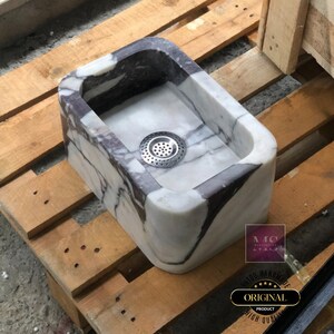 Hand-carved Calacatta Viola marble sink on a wooden pallet, showing the rectangular bowl, rounded corners and metal drain strainer.