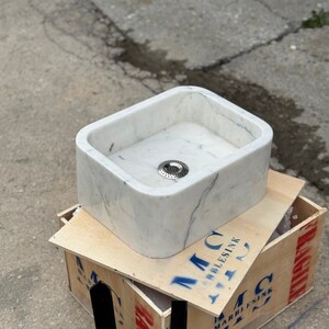 Right three-quarter workshop view of a Carrara White marble bathroom sink showing its thick solid stone walls, rounded corners, centered drain and black wall-mount brackets.
