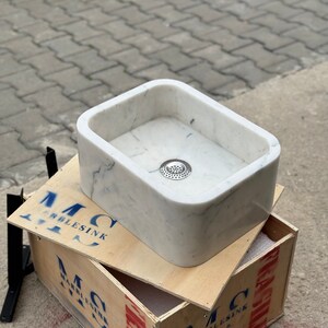 Front three-quarter view of a handmade Carrara White marble vessel sink with rounded rectangular corners, soft grey veining, deep polished bowl and centered metal drain.