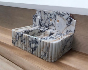 May include: A marble sink with a fluted front and a backsplash. The sink is white with black and gray veining. The backsplash has two circular holes. The sink is set against a light wood surface.