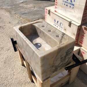 Right three-quarter workshop view of a rectangular Calacatta Viola marble sink, showing the polished natural stone, deep basin, centered metal drain, three faucet holes and heavy-duty wall brackets.