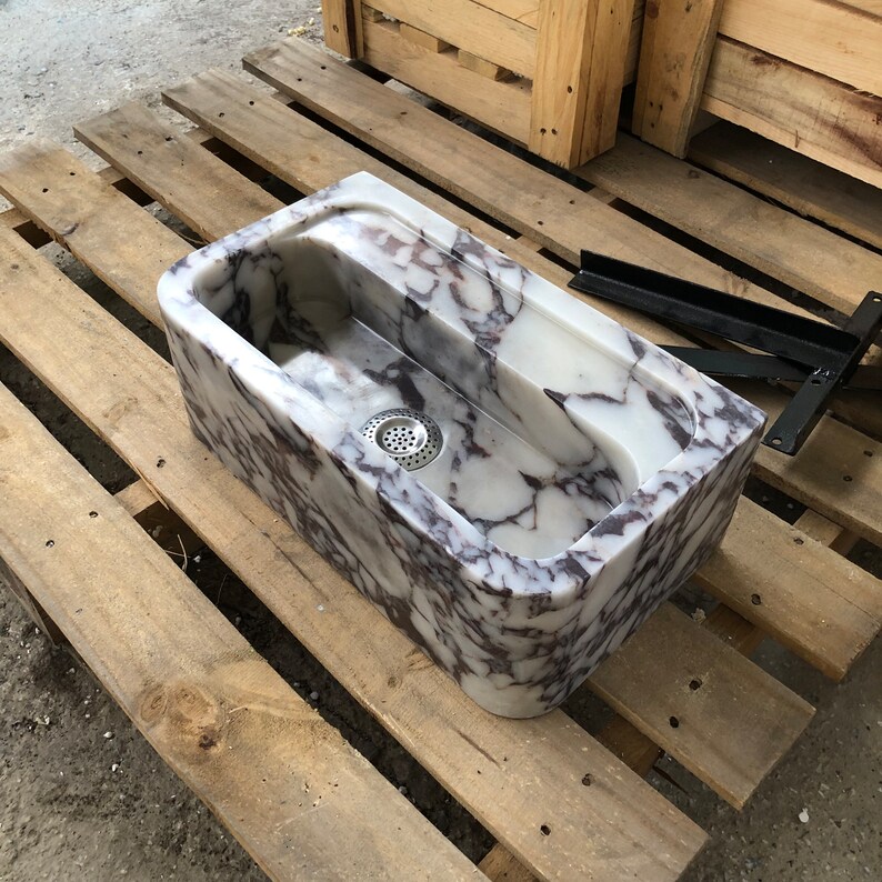 Op de afbeelding: Een witte marmeren gootsteen met paarse en grijze aderen. De gootsteen is rechthoekig en heeft een afgeronde rand. Hij staat op een houten pallet.
