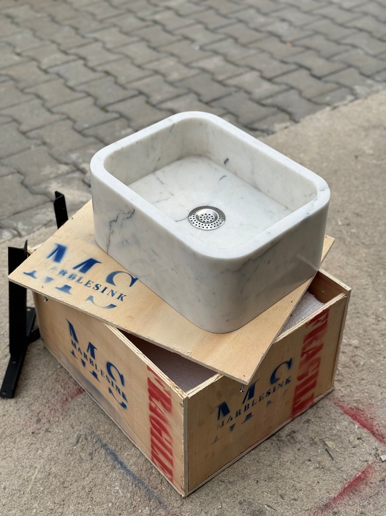 Front three-quarter view of a handmade Carrara White marble vessel sink with rounded rectangular corners, soft grey veining, deep polished bowl and centered metal drain.