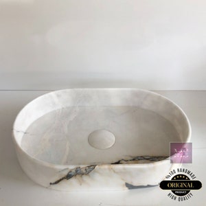 May include: White oval marble sink with a drain hole. The sink has a unique design with a slight curve on one side. The sink is handmade and has a high quality finish.