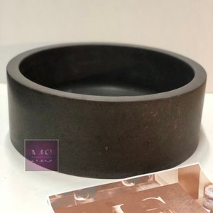 Pu&ograve; includere: Un vaso rotondo in cemento grigio scuro con una superficie liscia e leggermente ruvida.
