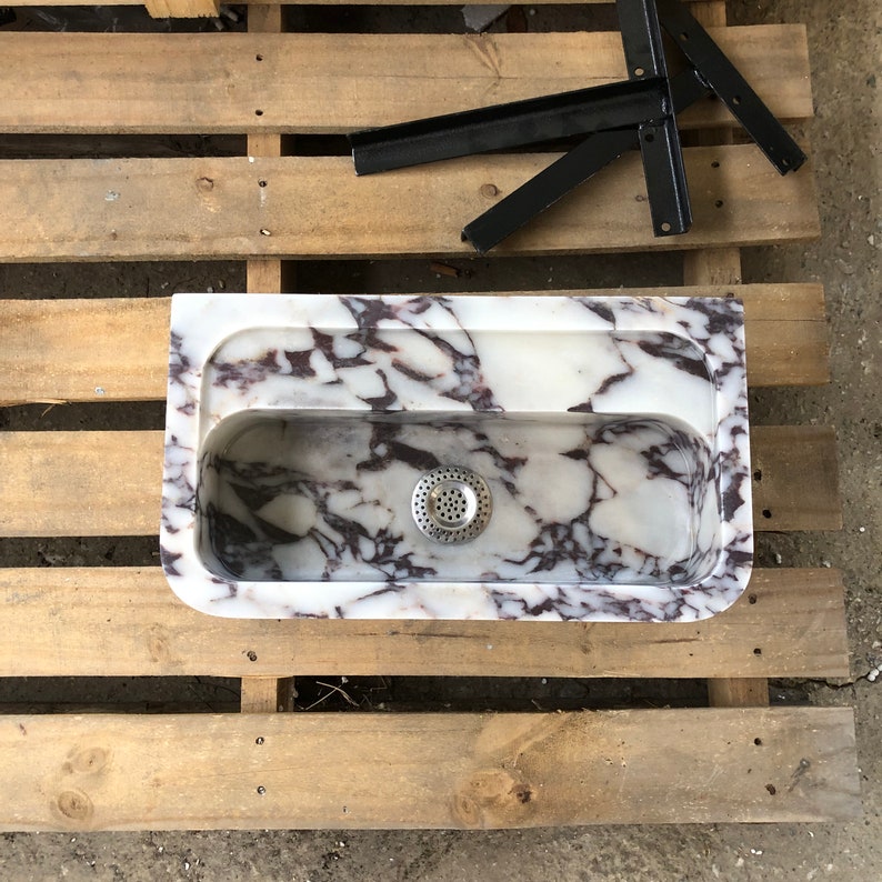 Op de afbeelding: Een witte marmeren gootsteen met paarse aderen. De gootsteen heeft &eacute;&eacute;n afvoer en staat op een houten pallet.