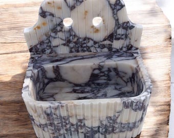 May include: A white marble soap dish with a decorative back and two holes. The dish has a rectangular shape and a ribbed design.