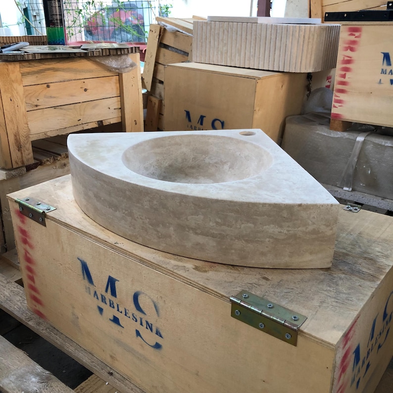May include: A corner sink made of travertine stone. The sink is in a wooden crate with the text "MC Marblesink" printed on the side.