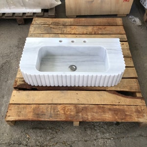 May include: White marble kitchen sink with a fluted edge and three holes for taps. The sink is sitting on a wooden pallet.