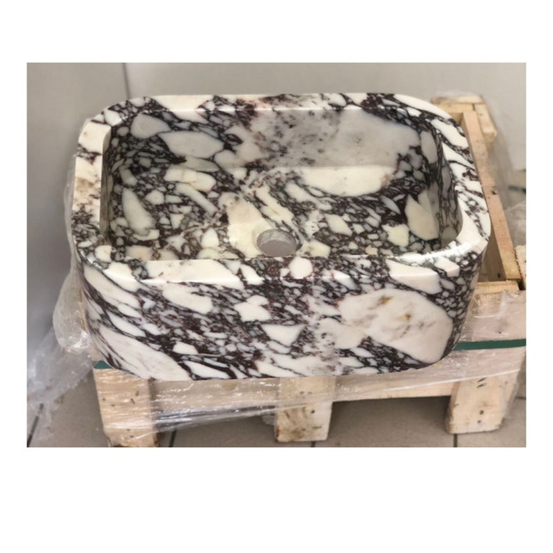 May include: A white marble sink with purple and brown veining. The sink is rectangular with rounded edges and has a single drain hole.