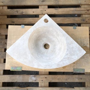 May include: A white corner sink made of natural stone with a round basin and a single drain hole. The sink is mounted on a wooden surface.