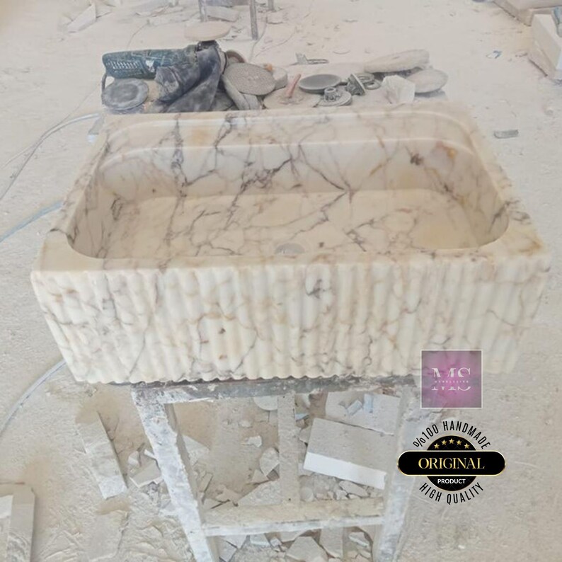May include: A white marble sink with a rippled edge. The sink is handmade and has a 100% original product guarantee. The sink is on a wooden stand.