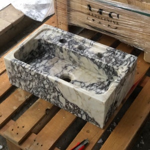Puede incluir: Un lavabo de m&aacute;rmol blanco con vetas moradas y doradas. El lavabo tiene cuatro agujeros para grifos y un agujero de drenaje en la parte inferior. El lavabo est&aacute; sentado en una paleta de madera.