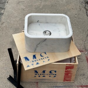 Front overhead view of a handmade Carrara marble washbasin with a deep polished bowl, centered metal drain, rounded inner corners and natural white and grey stone pattern.
