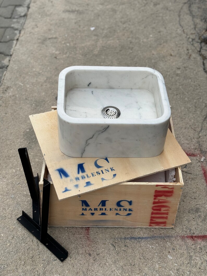Front overhead view of a handmade Carrara marble washbasin with a deep polished bowl, centered metal drain, rounded inner corners and natural white and grey stone pattern.