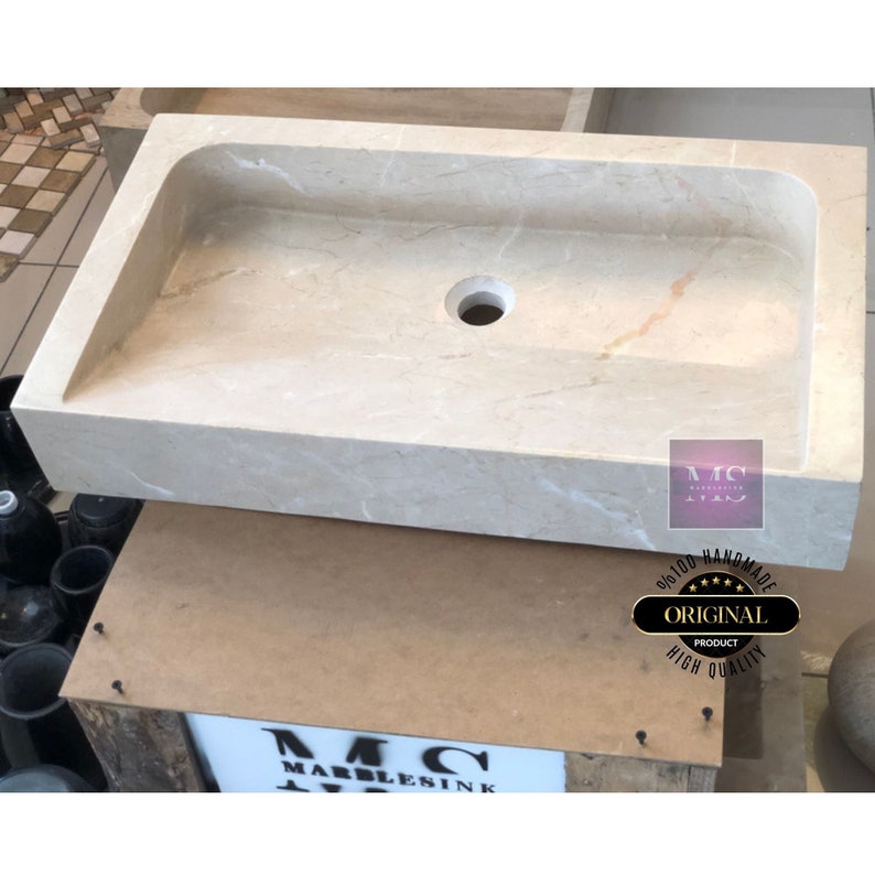 May include: A rectangular, off-white marble sink with a single drain hole. The sink is handmade and features a subtle veining pattern. The sink is sitting on a wooden surface with a label that reads "MC MARBLESINK".