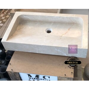 May include: A rectangular, off-white marble sink with a single drain hole. The sink is handmade and has a smooth, polished finish. The sink is sitting on a wooden surface with a label that reads "100% Handmade Original Product High Quality".